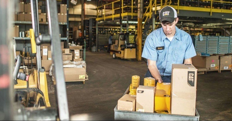 A warehouse worker wearing a blue shirt and a CAT baseball cap stands behind a utility cart filled with boxed CAT parts and yellow filters. In the background, an industrial warehouse setting features shelves of inventory, cardboard boxes, and a distant forklift.